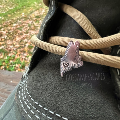 Solid copper shoelace charm in the shape of the state of Maine with a heart stamped into the lower left side.  The edges of the charm have a hammered texture.  The charm is installed on a boot shoelace.  Fall leaves are on the ground in the background.